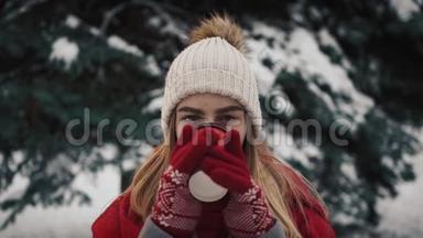 年轻漂亮的女孩穿着暖和的衣服站在雪中的圣诞树旁边，从纸杯里喝咖啡